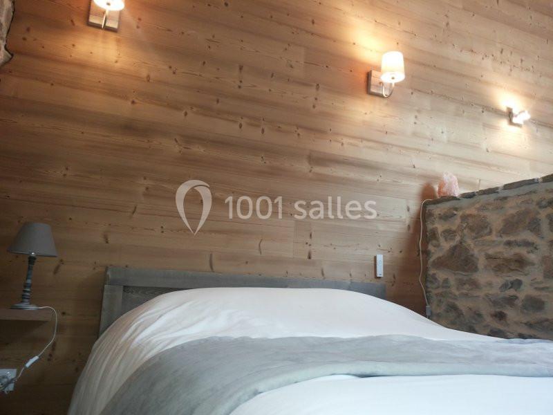 Lit avec draps blancs et couverture grise dans une chambre aux murs en bois et pierre, éclairée par des appliques murales.