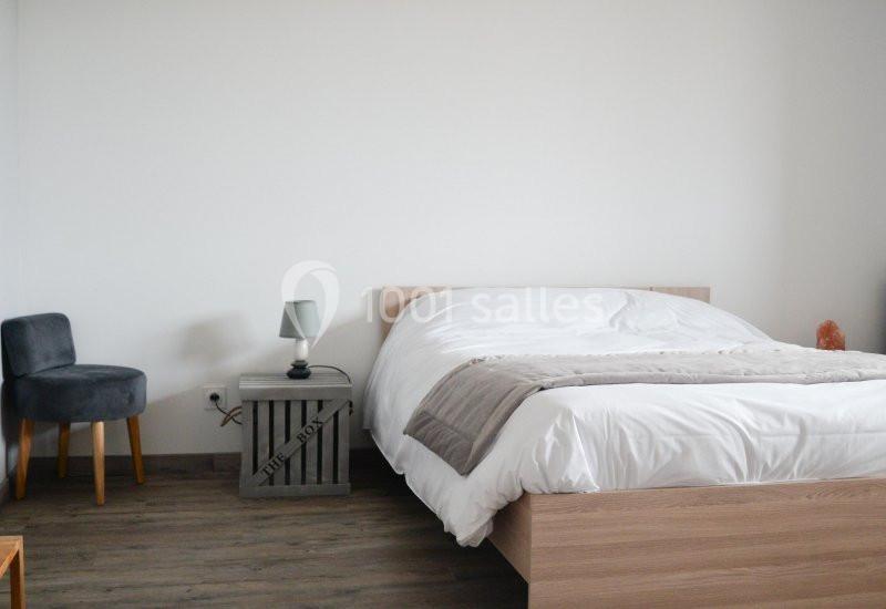 Chambre minimaliste avec un lit double, une table de chevet en bois, une lampe et une chaise grise sur un sol en parquet.