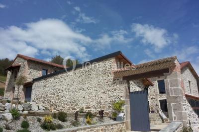 Façade en pierre d'une maison rénovée avec terrasse, grandes fenêtres et espace extérieur gravillonné.