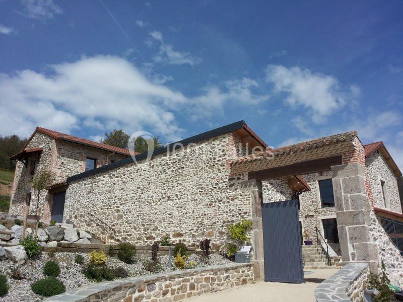 Bâtisse en pierre rénovée avec toit en tuiles, entourée d'un jardin paysager sous un ciel partiellement nuageux.