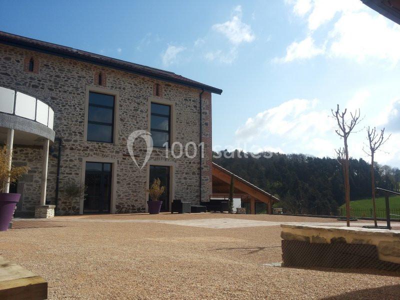 Façade en pierre d'une maison moderne avec terrasse en gravier, arbres taillés et vue sur la campagne.