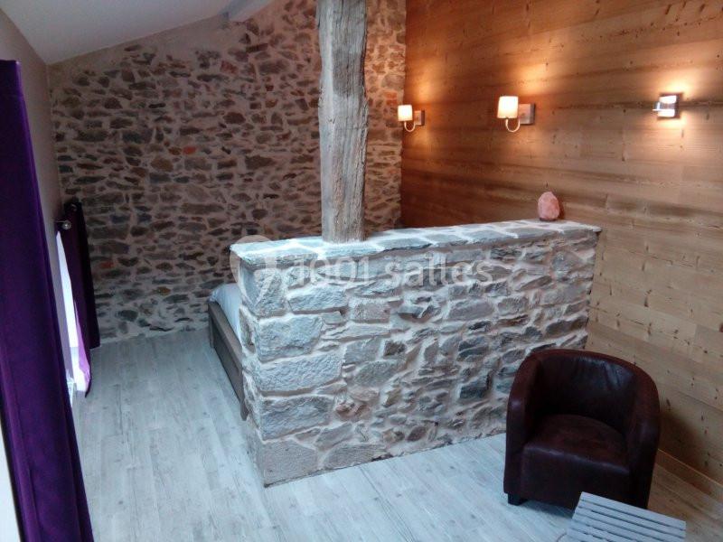 Mur en pierre apparente avec poutre en bois, fauteuil en cuir et éclairage mural dans une pièce au style rustique.