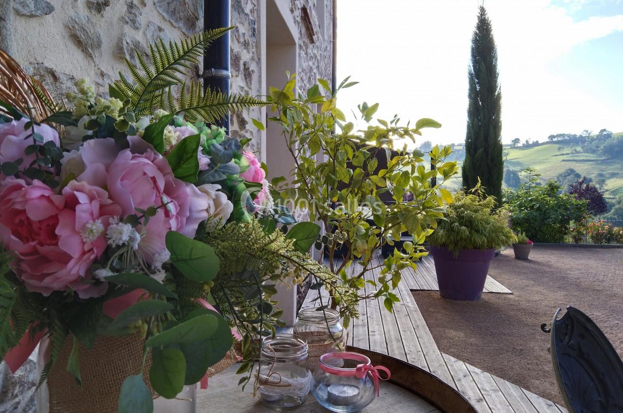Bouquet de fleurs roses et feuillages sur une table en bois, avec vue sur un jardin et un paysage vallonné en arrière-plan.
