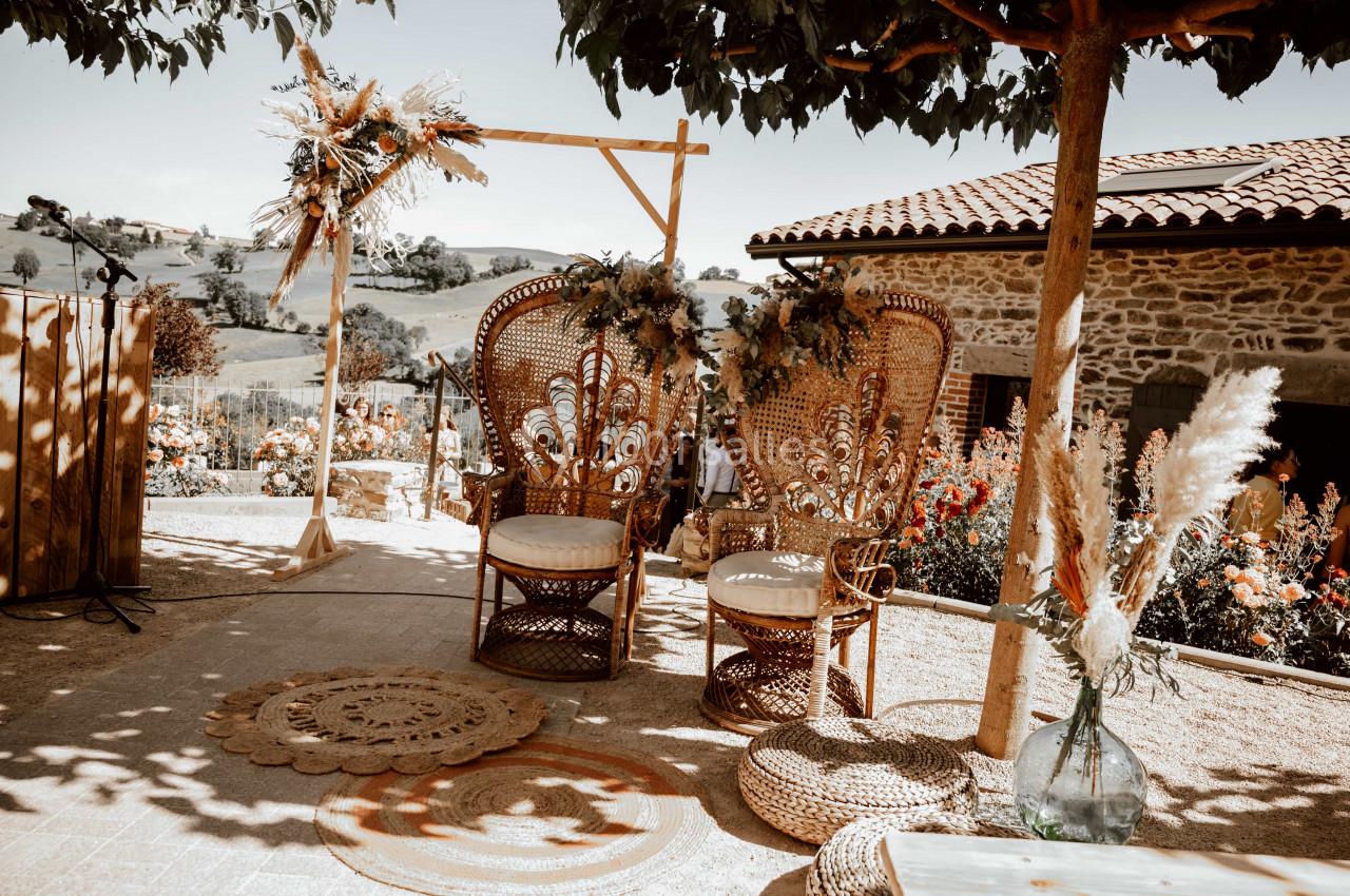 Deux fauteuils en rotin décorés de fleurs, installés dans un espace extérieur ensoleillé avec vue sur la campagne.