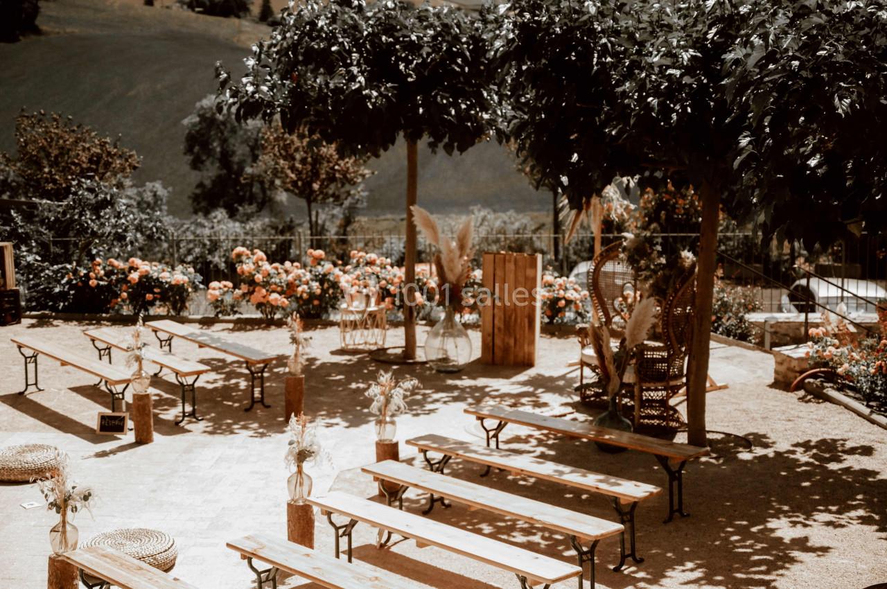 Espace extérieur aménagé avec bancs en bois, décorations florales et arbres offrant de l'ombre.