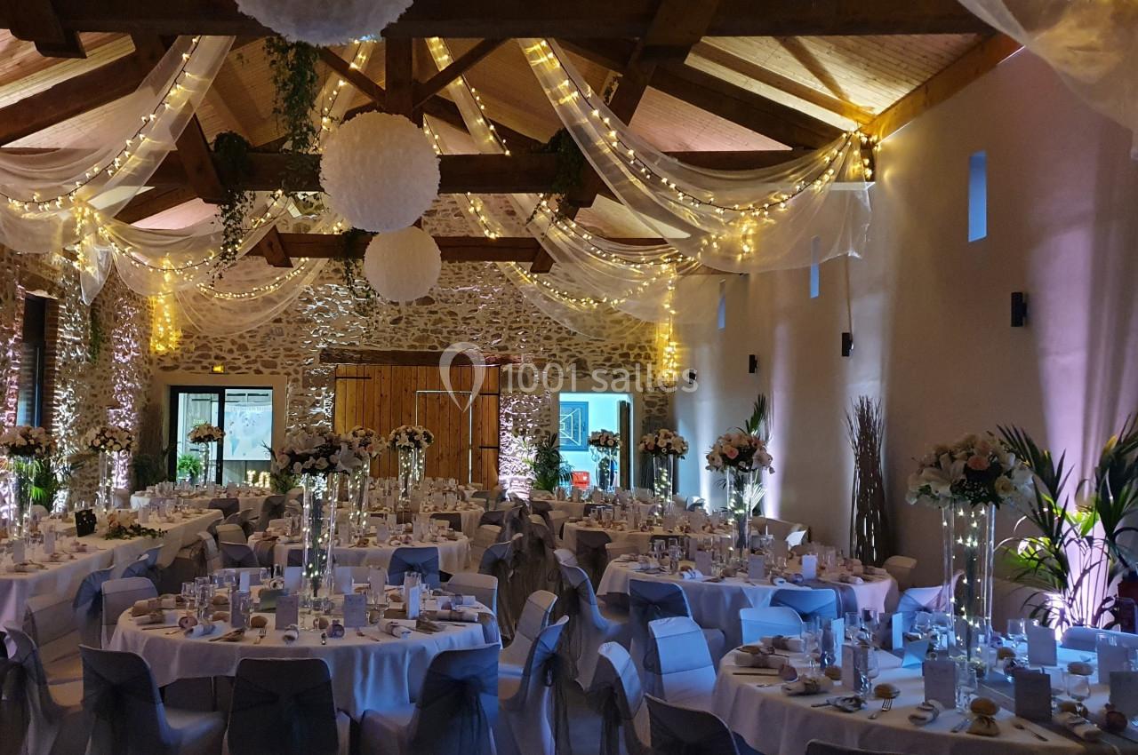 Salle de réception décorée pour un mariage, avec tables rondes dressées, guirlandes lumineuses et voilages suspendus.