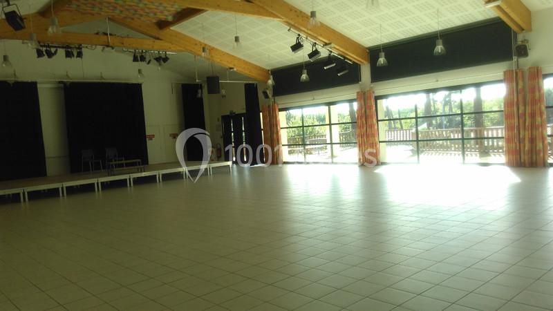 Salle polyvalente lumineuse avec sol carrelé, scène à gauche, grandes baies vitrées et poutres apparentes au plafond.