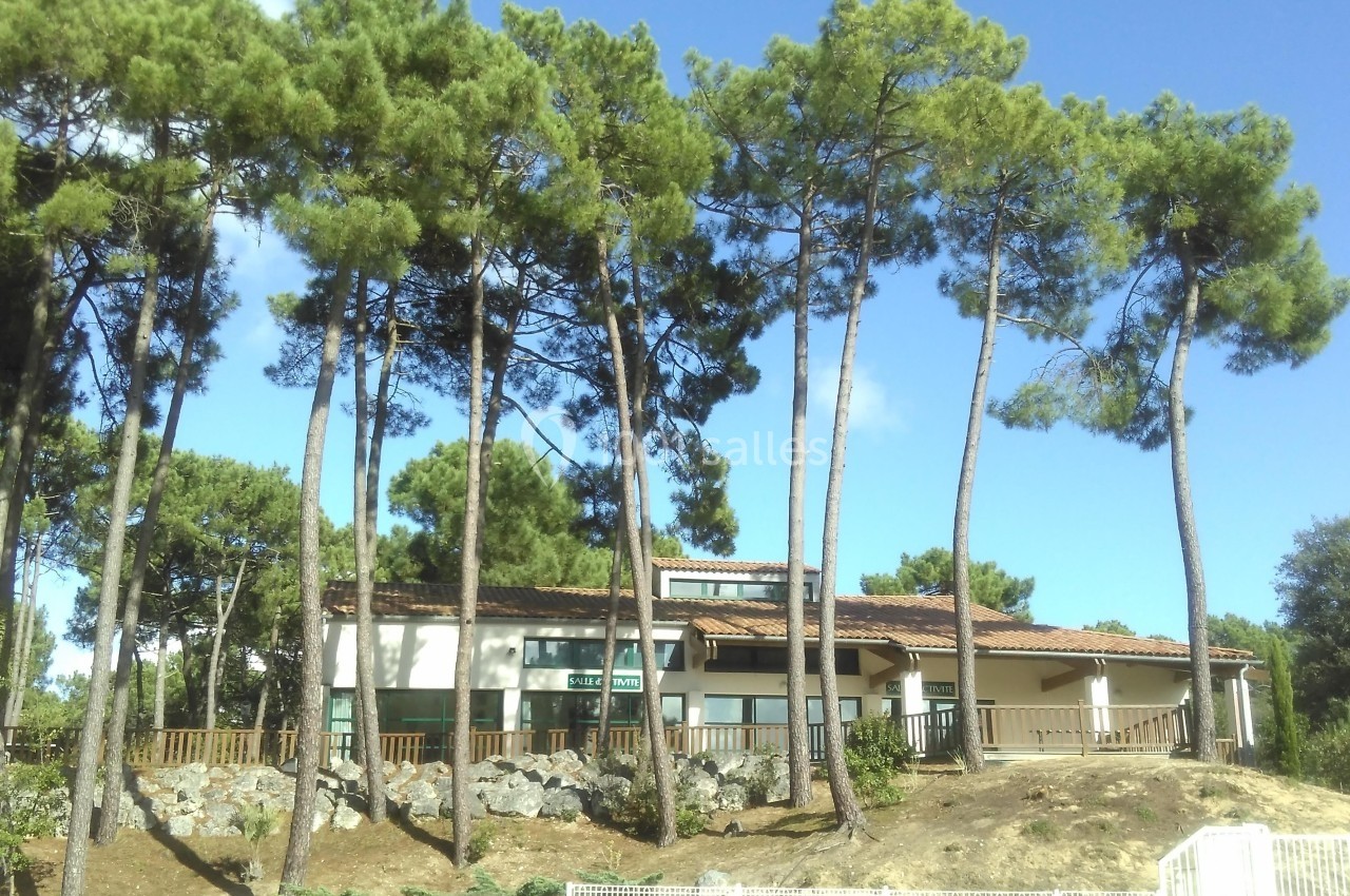 Bâtiment entouré de pins sous un ciel bleu, avec une clôture et un terrain légèrement en pente au premier plan.