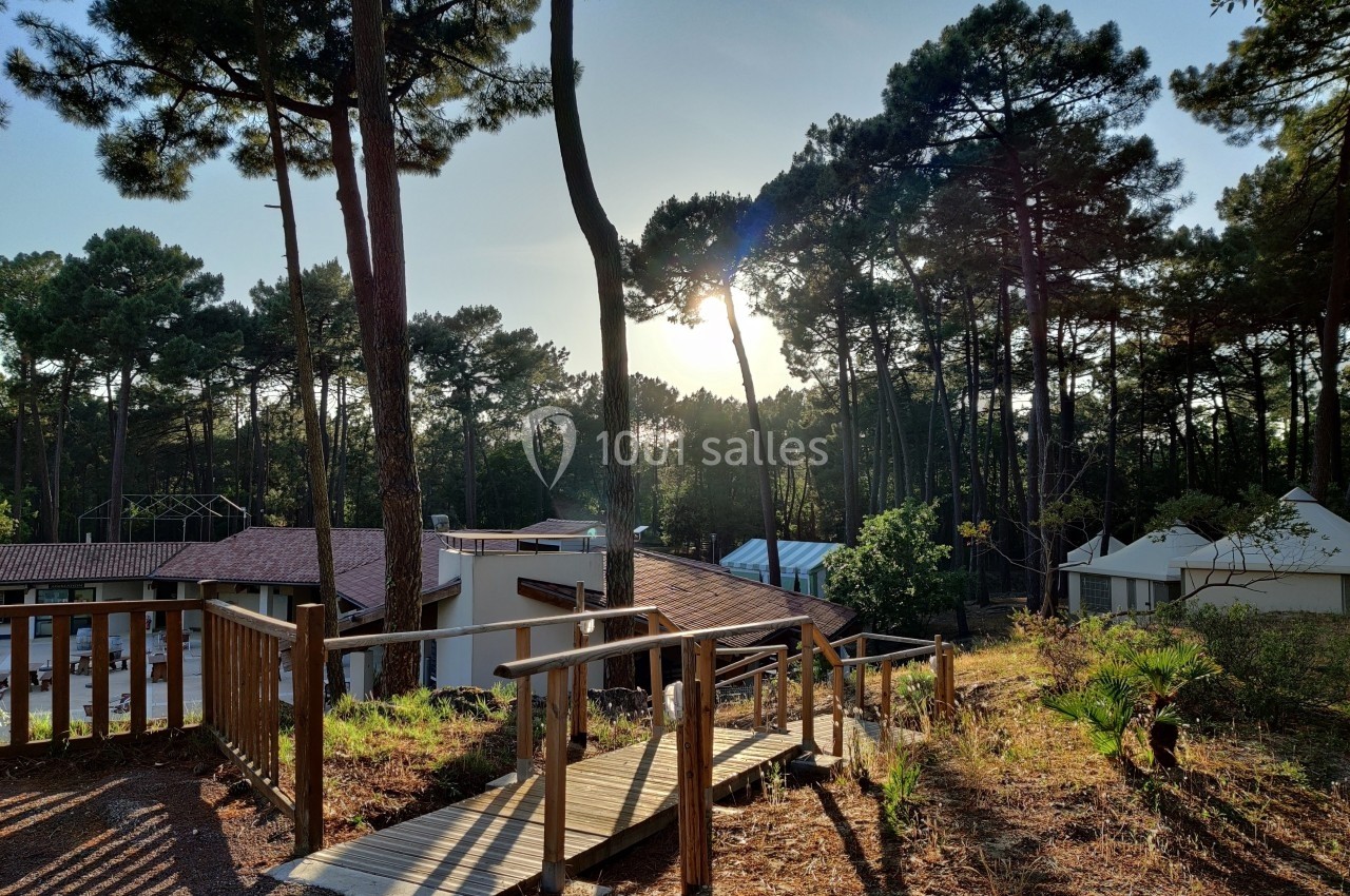 Passerelle en bois entourée de pins, menant à des bâtiments sous un ciel ensoleillé en fin de journée.