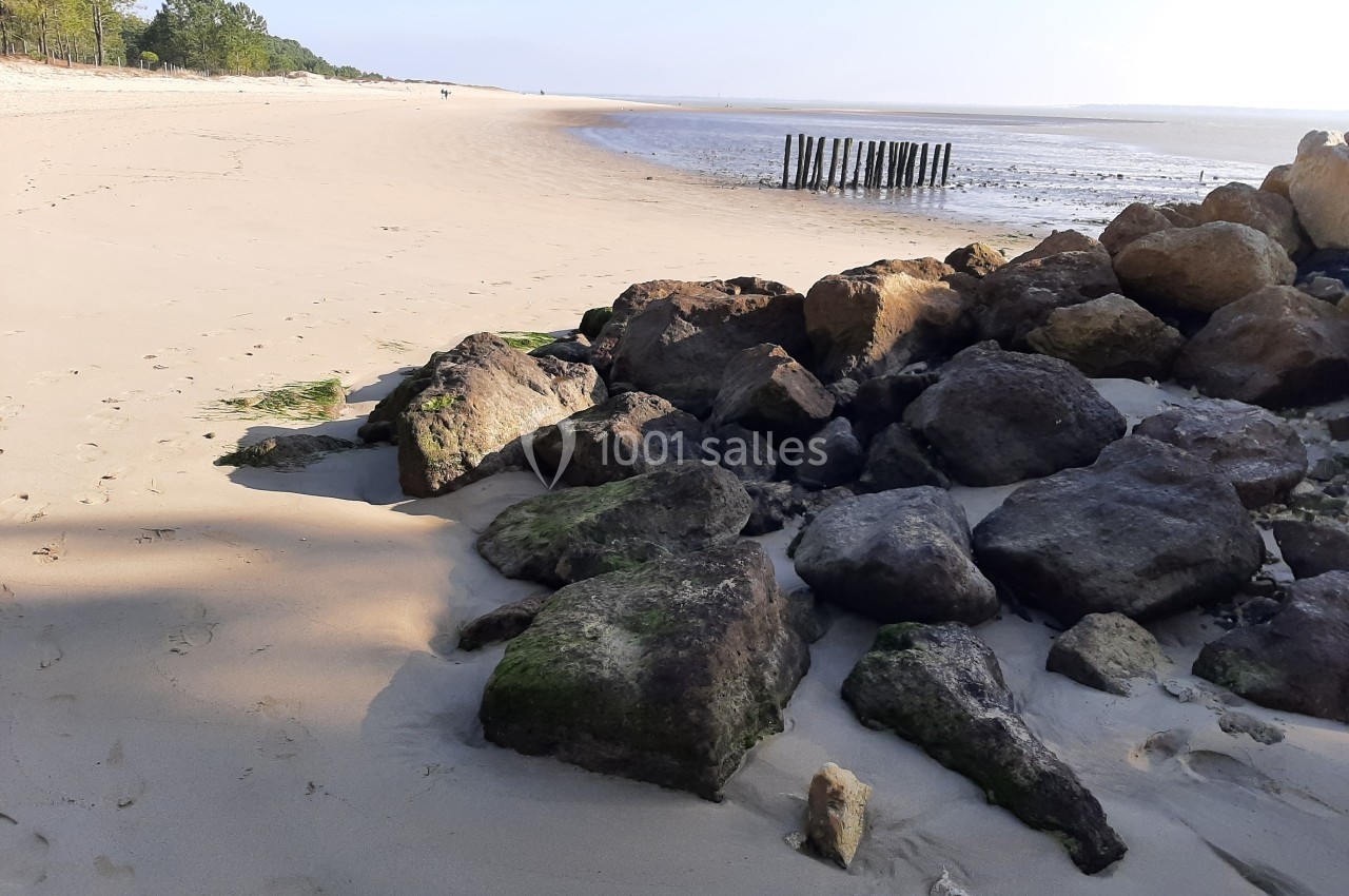 Plage de sable avec des rochers au premier plan et des poteaux en bois enfoncés dans le sol près de l'eau.
