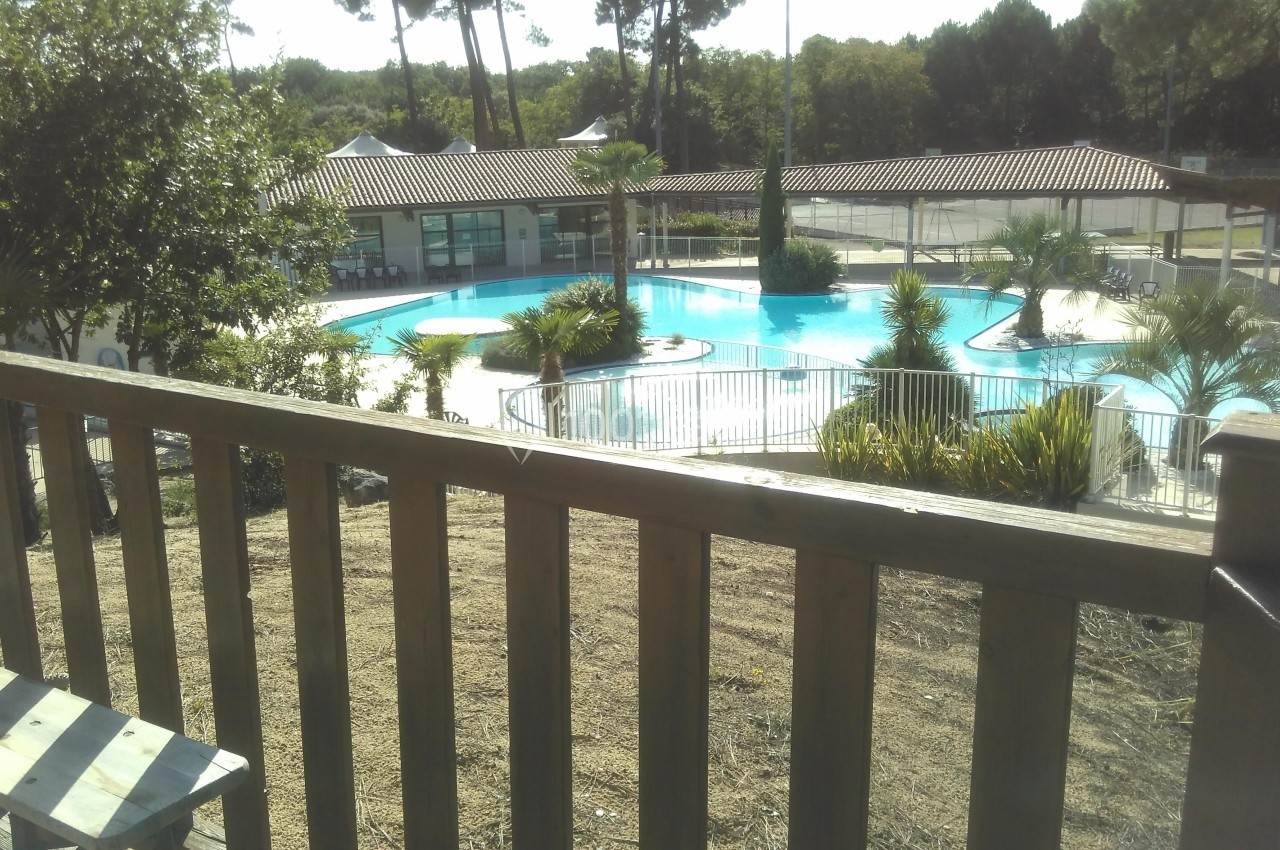 Vue d'une piscine extérieure entourée de végétation, avec des bâtiments en arrière-plan et une barrière au premier plan.