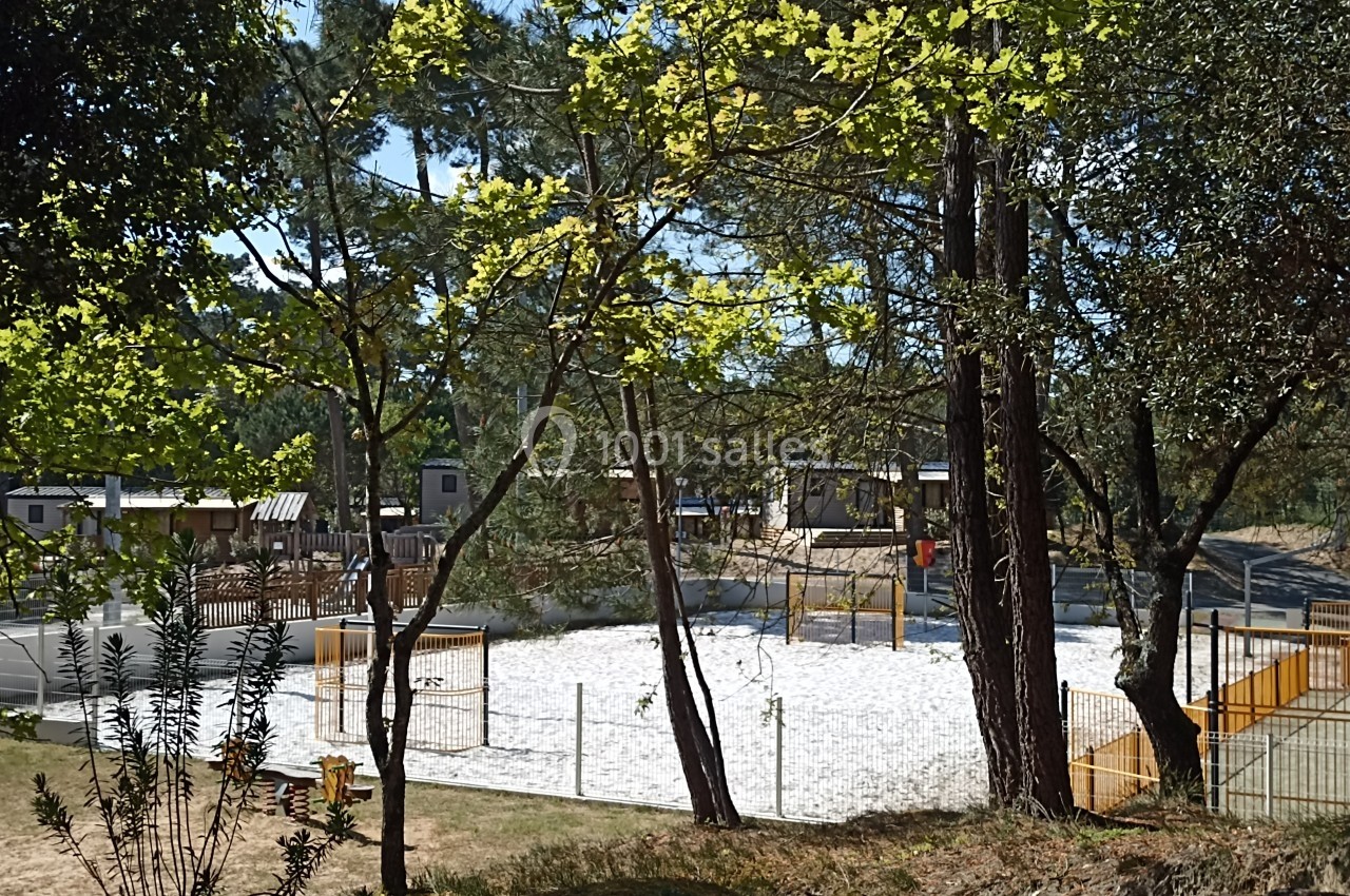 Terrain de volley en sable entouré de clôtures, situé dans un espace boisé avec des mobil-homes en arrière-plan.