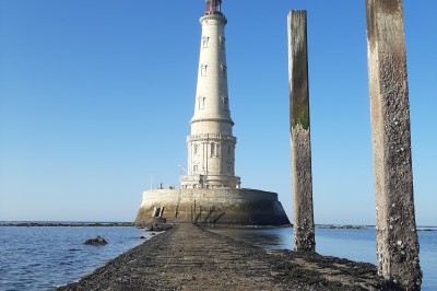 Phare blanc sur un îlot rocheux, accessible par une jetée étroite, entouré d'eau sous un ciel bleu clair.