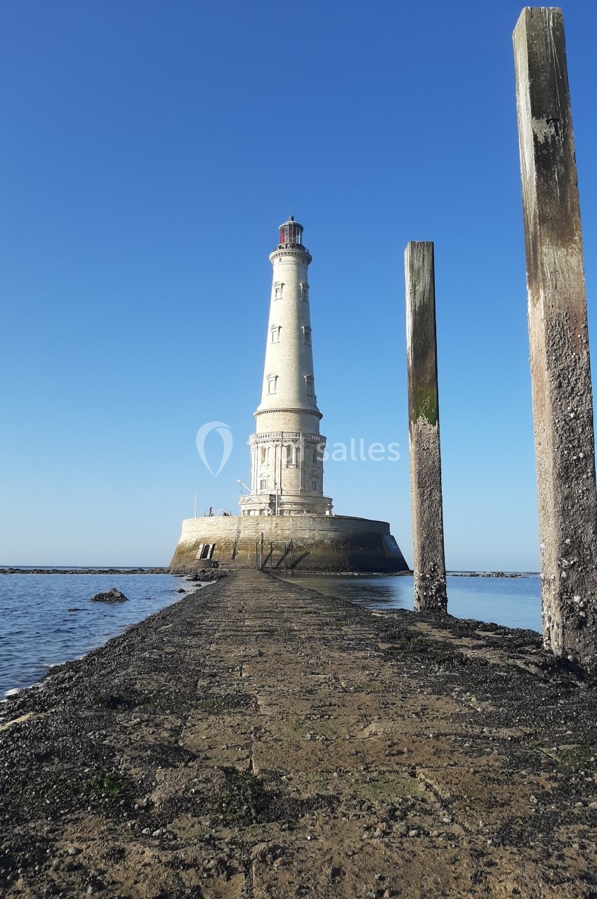 Phare blanc sur un îlot rocheux, accessible par une jetée étroite, entouré d'eau sous un ciel bleu clair.