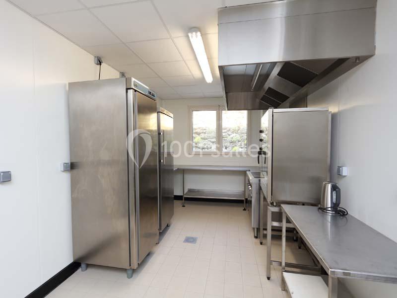 Cuisine professionnelle équipée d'appareils en inox, avec réfrigérateur, hotte, plan de travail et éclairage fluorescent.