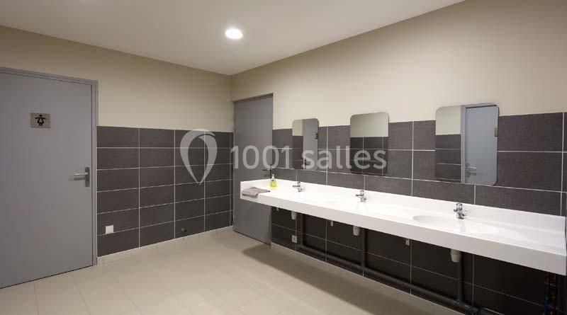 Salle de bain publique avec trois lavabos, trois miroirs et des murs carrelés gris.