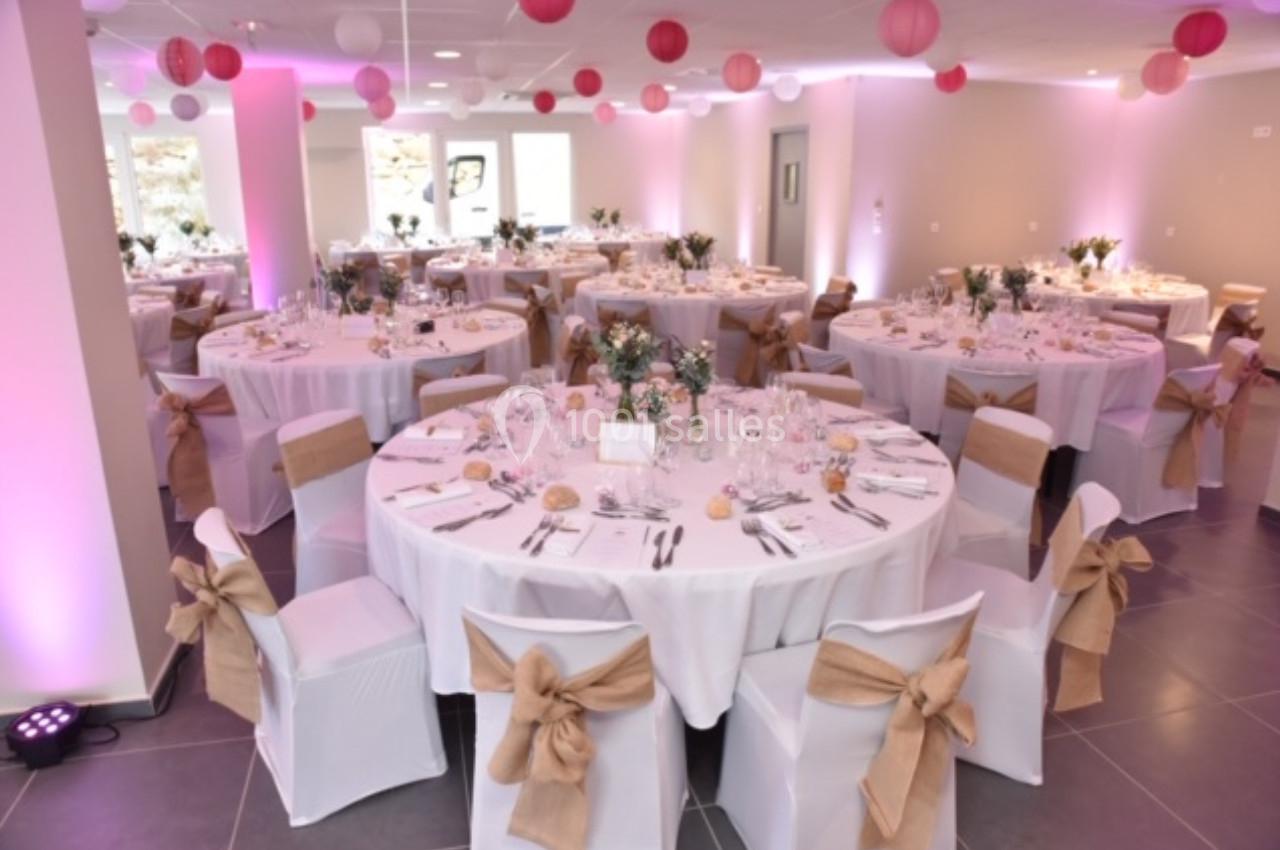 Salle de réception décorée avec des tables rondes, nappes blanches, chaises ornées de nœuds et éclairage rose.