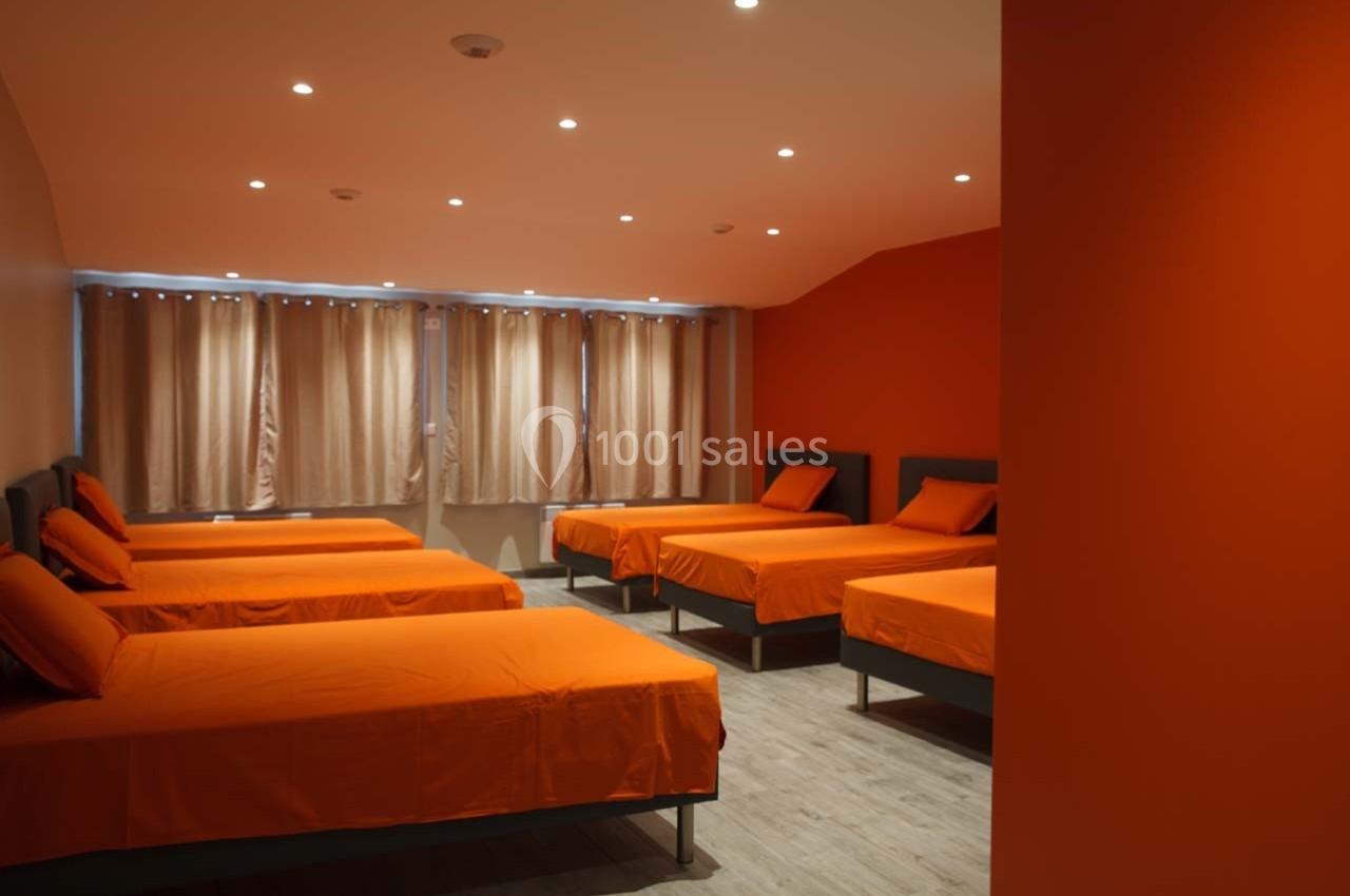 Dortoir avec plusieurs lits simples aux draps orange, éclairé par des spots au plafond et une fenêtre avec rideaux beiges.
