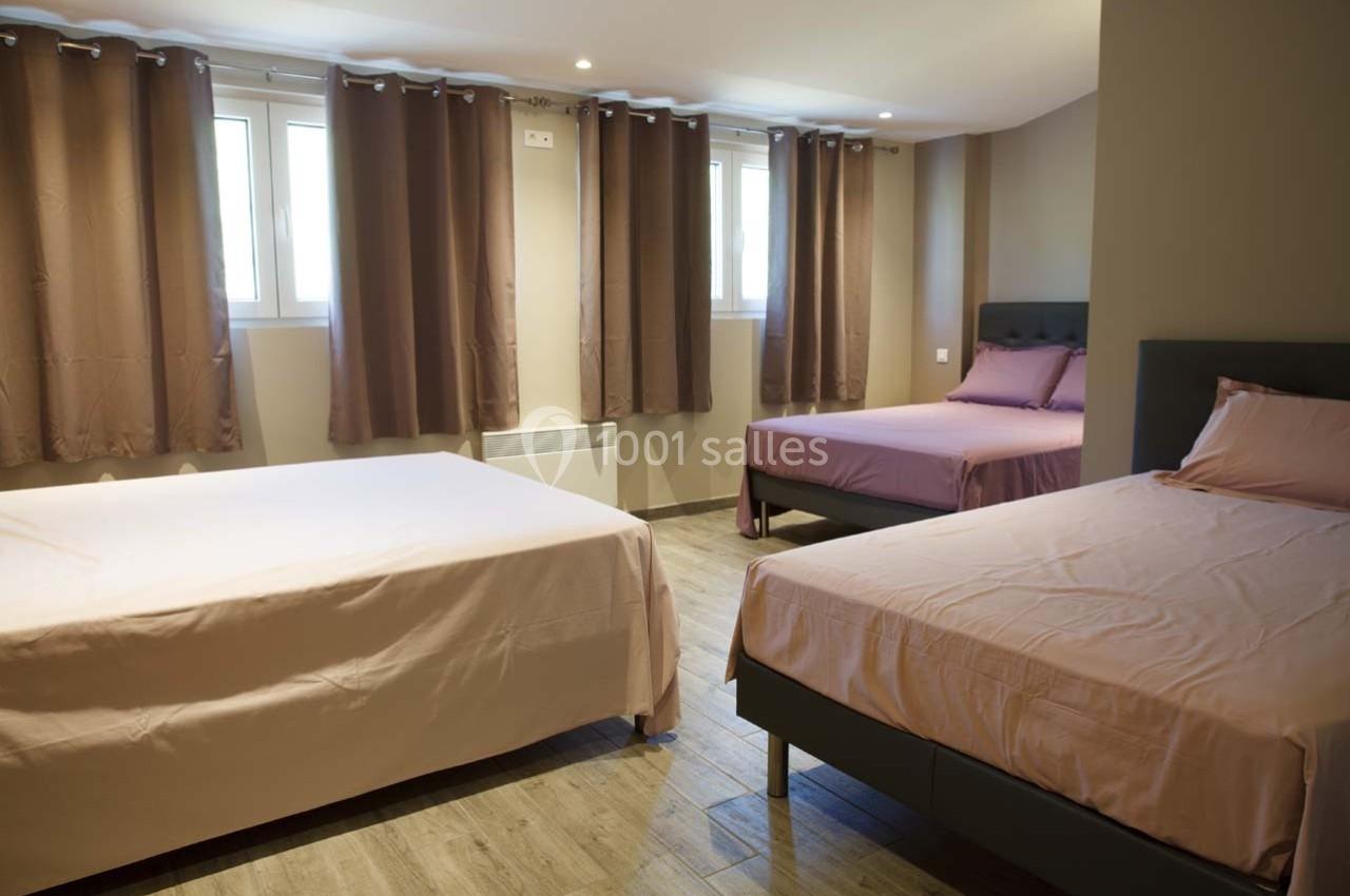 Chambre avec trois lits doubles, parquet au sol, murs beiges et rideaux marron couvrant des fenêtres.