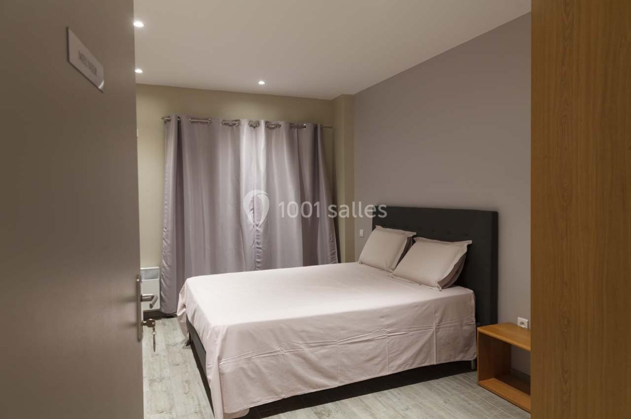 Chambre avec lit double, draps clairs, rideaux gris, sol en parquet et éclairage encastré.