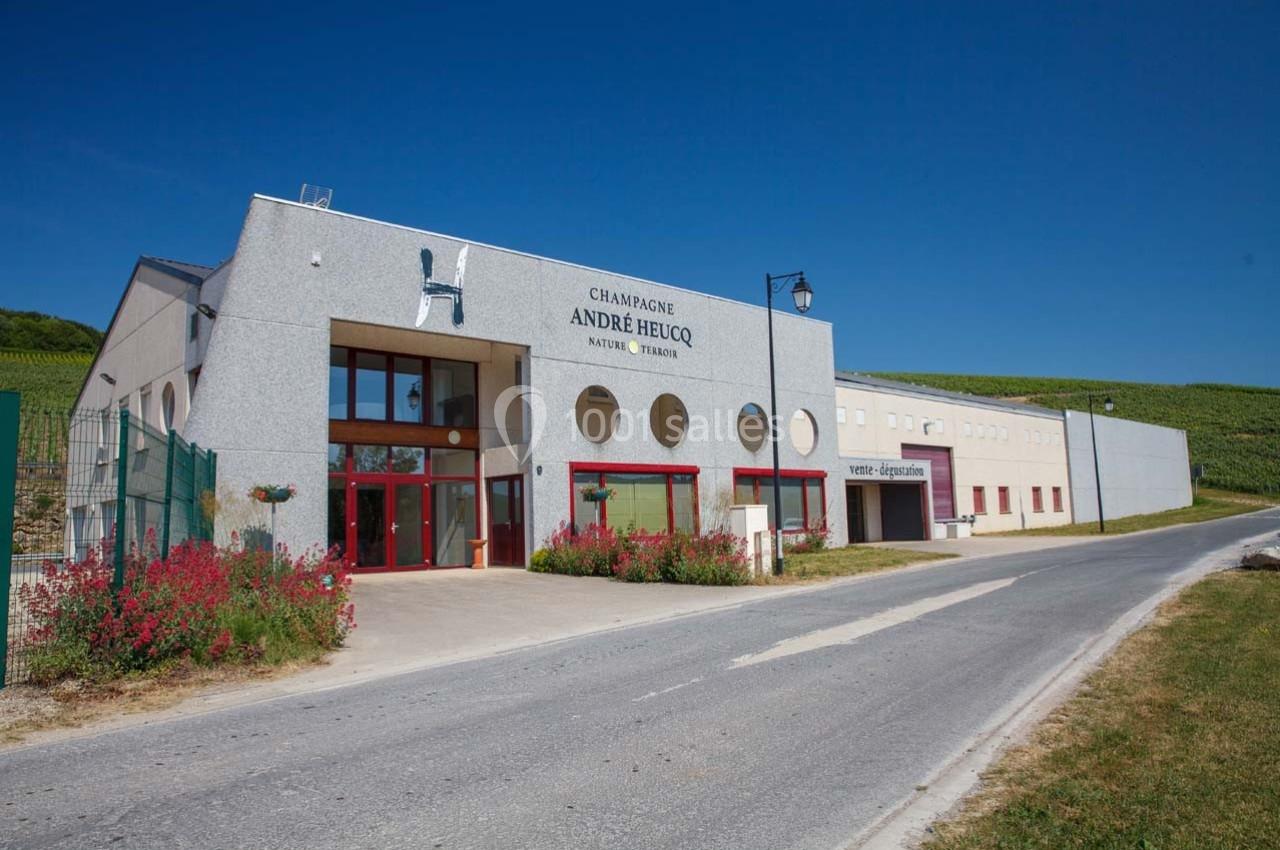 Bâtiment moderne du domaine viticole André Heucq, entouré de vignes, avec une route adjacente.