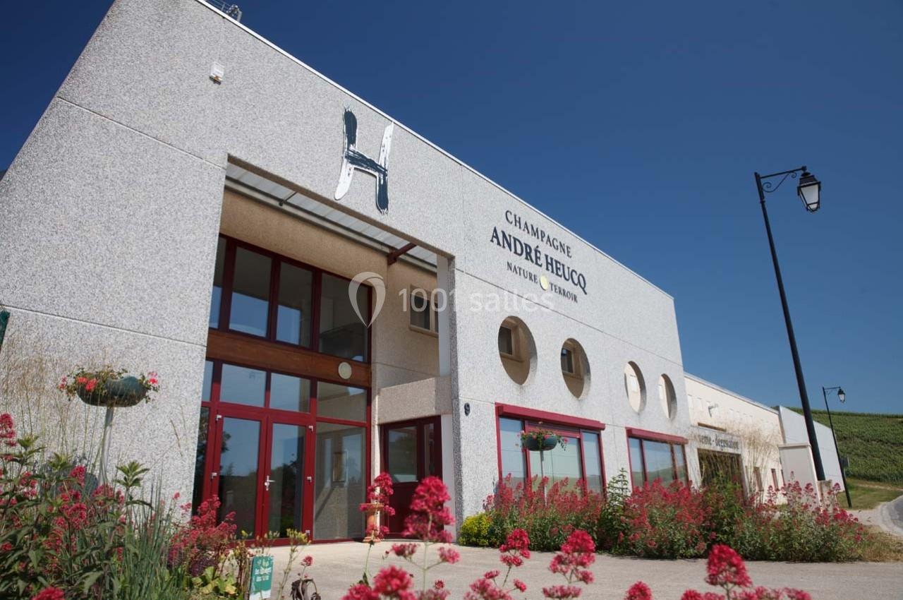 Façade d'un bâtiment moderne avec enseigne ’Champagne André Heucq’, entouré de fleurs et d'un ciel bleu.