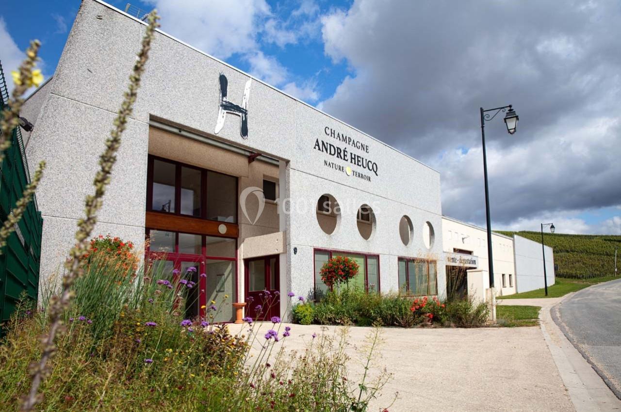 Façade d'un bâtiment viticole avec l'inscription ’Champagne André Heucq’, entouré de fleurs et de vignes.