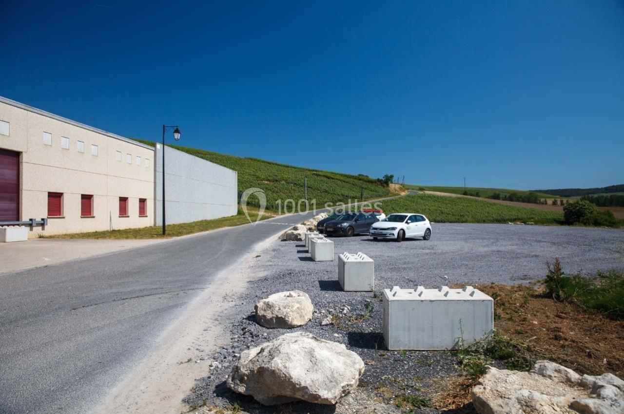Parking avec quelques voitures stationnées près d'un bâtiment industriel, entouré de champs et d'un ciel dégagé.