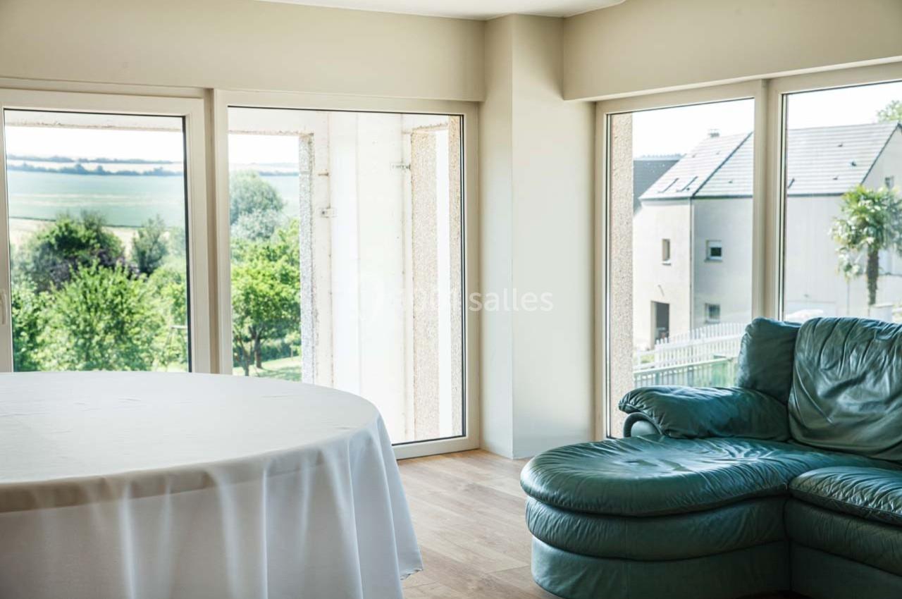 Salon lumineux avec grandes baies vitrées donnant sur un paysage verdoyant, canapé en cuir et table ronde.