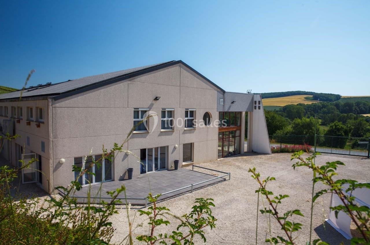 Bâtiment moderne avec façade beige, grandes baies vitrées et terrasse, entouré de végétation et d'un paysage rural.