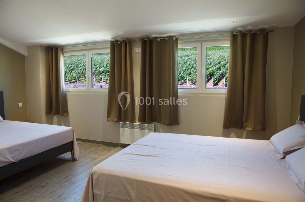 Chambre lumineuse avec deux lits, parquet au sol, murs clairs et fenêtres donnant sur un jardin verdoyant.