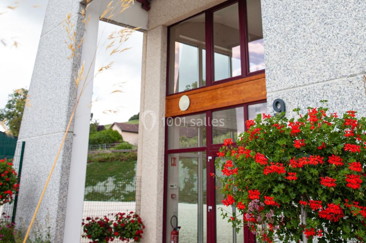 Entrée vitrée d'un bâtiment moderne avec des murs en pierre, entourée de jardinières de géraniums rouges.