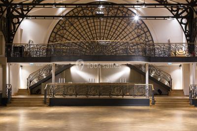Salle intérieure avec mezzanine, escaliers symétriques, balustrades en métal décoratif et parquet au sol.