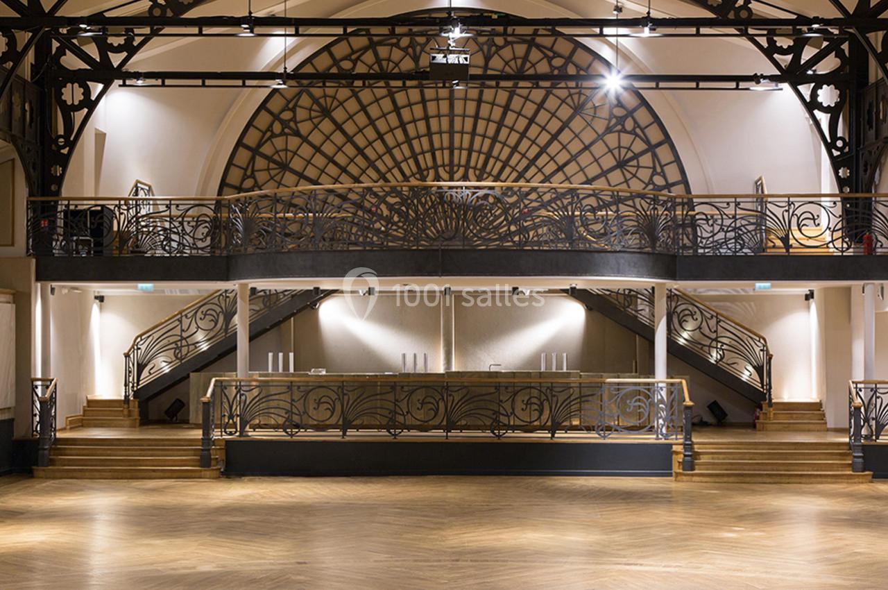 Salle intérieure avec mezzanine, escaliers symétriques, balustrades en métal décoratif et parquet au sol.
