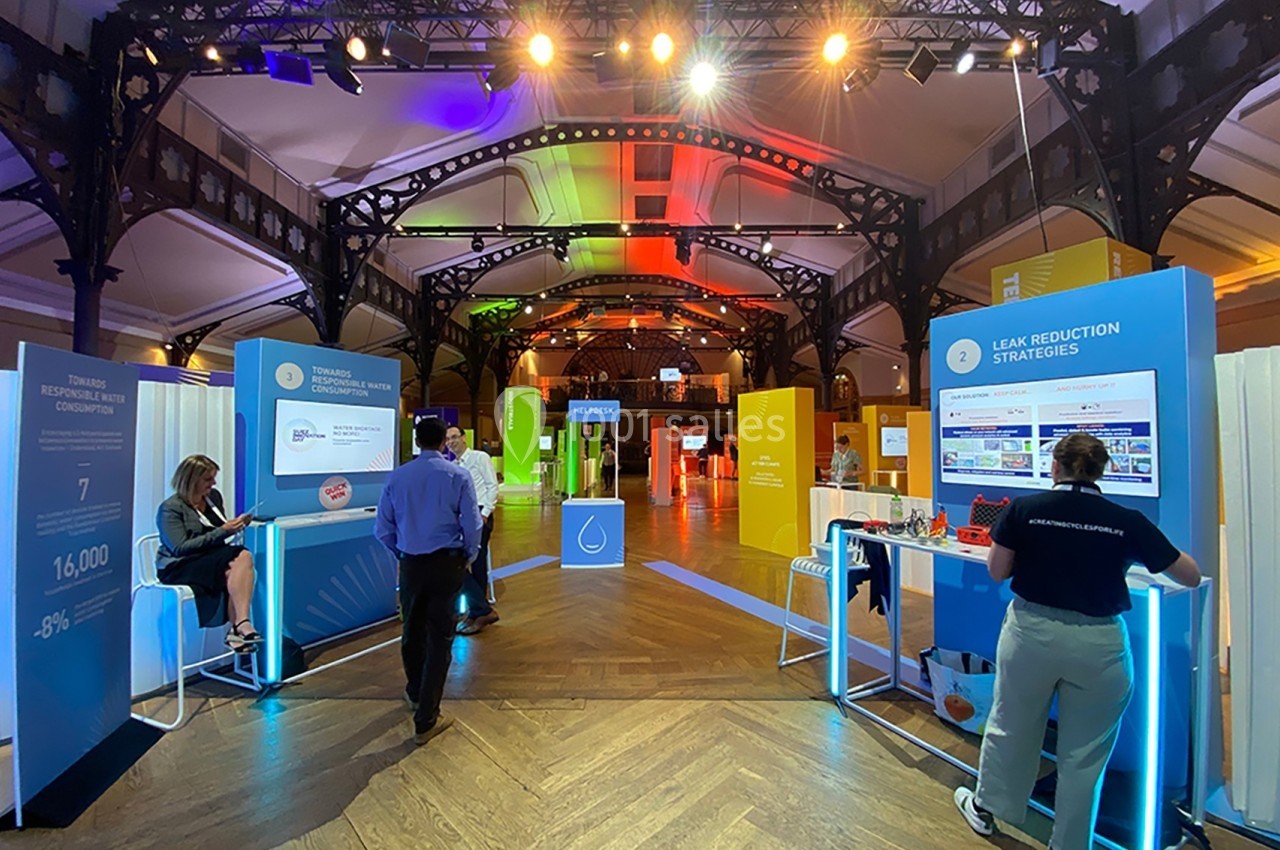 Salle d'exposition lumineuse avec stands interactifs, visiteurs debout et écrans présentant des stratégies de réduction des…
