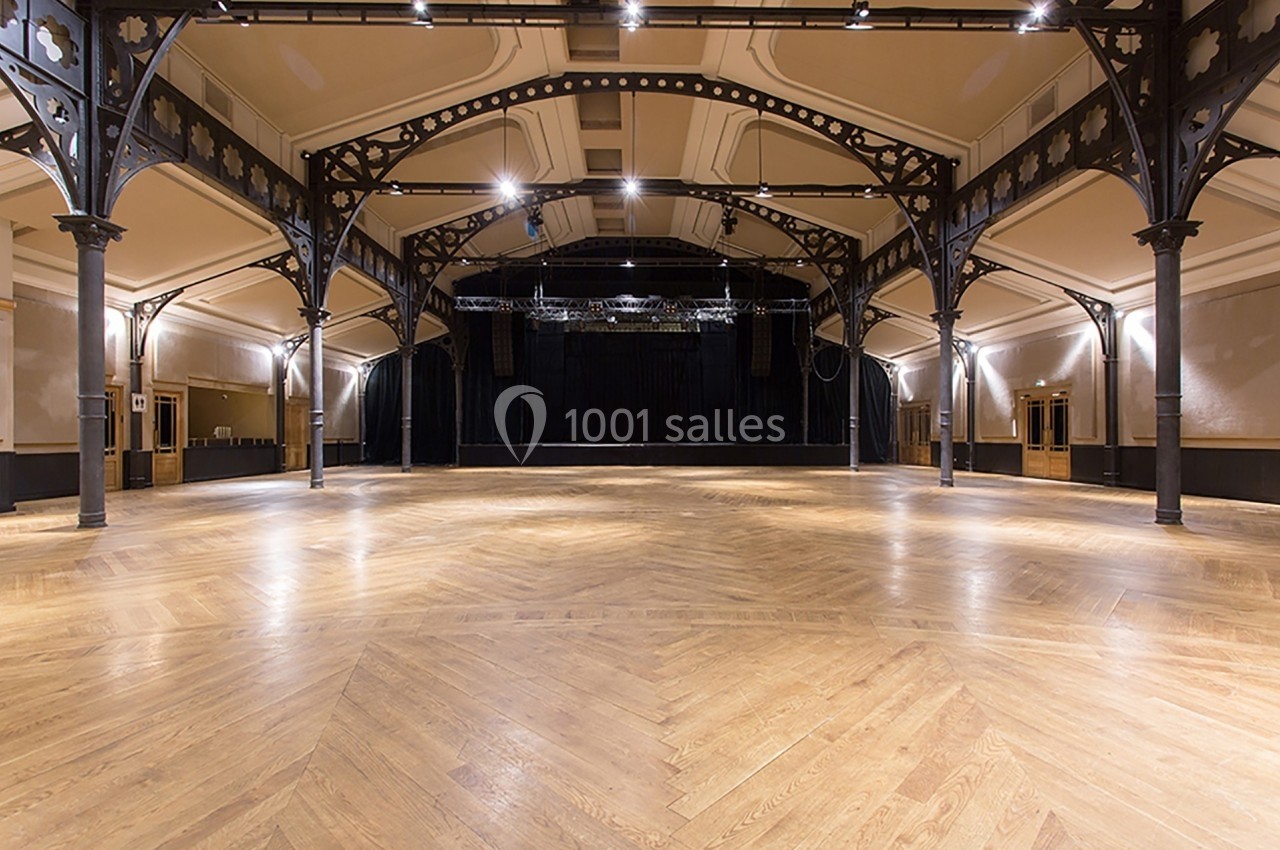 Salle spacieuse avec parquet en bois, poutres métalliques apparentes et scène noire en fond.