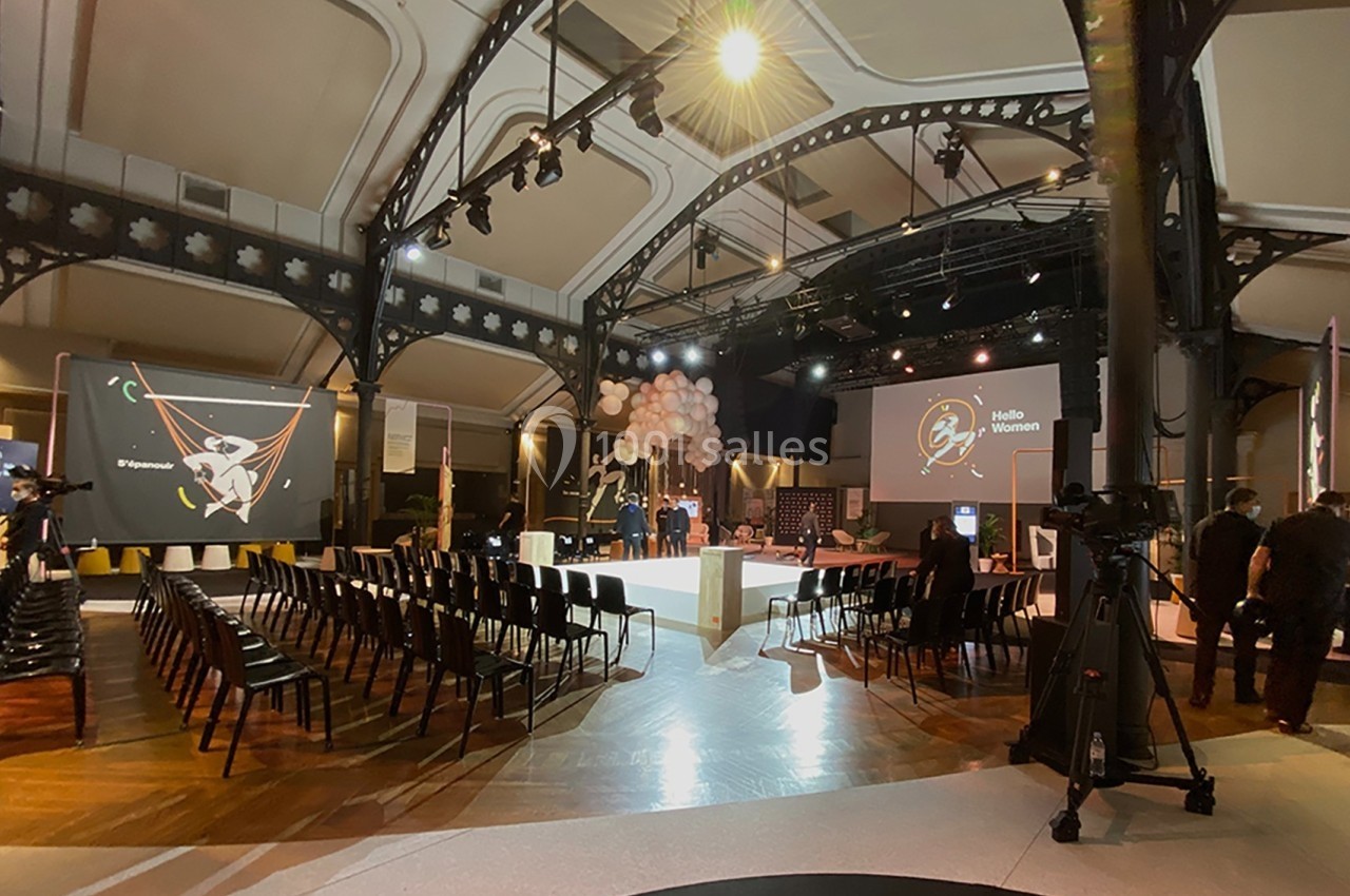 Salle de conférence avec des rangées de chaises vides, une scène éclairée et des écrans affichant des visuels.