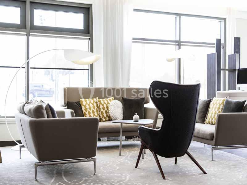 Salon lumineux avec canapés beiges, fauteuil noir, coussins décoratifs et lampadaire arqué près de grandes fenêtres.