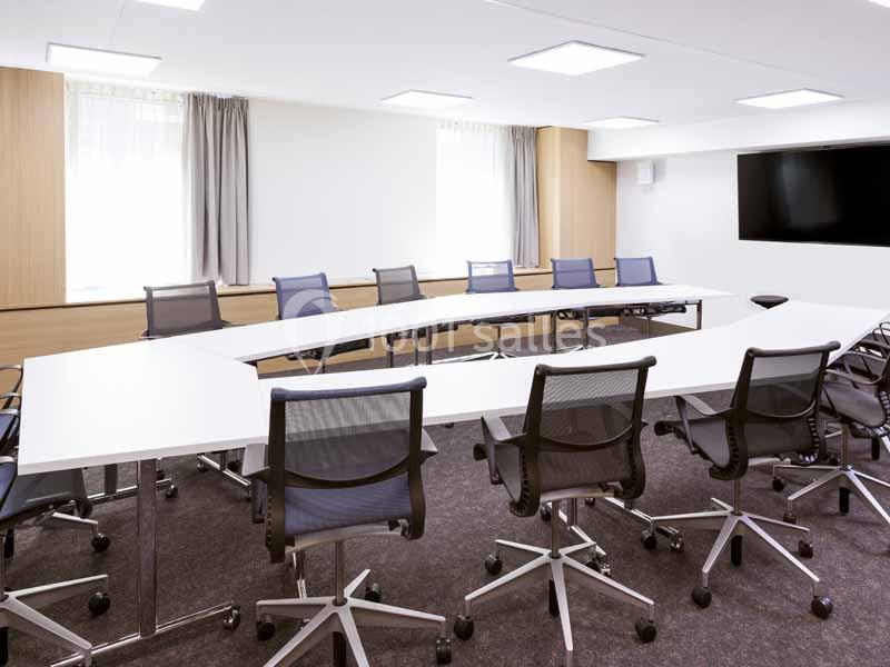 Salle de réunion lumineuse avec une grande table en U, des chaises à roulettes et un écran mural.