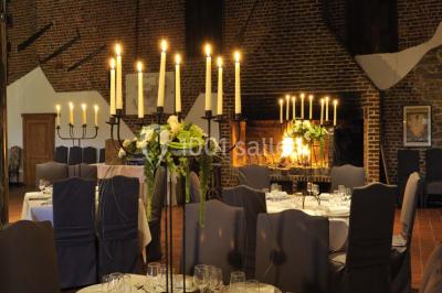 Salle de réception avec tables dressées, nappes beiges, chandeliers et poutres apparentes sous un toit vitré.