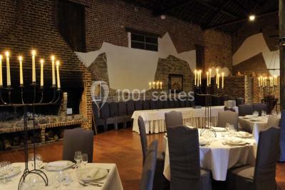 Salle de réception avec tables dressées, nappes beiges, chandeliers et poutres apparentes sous un toit vitré.