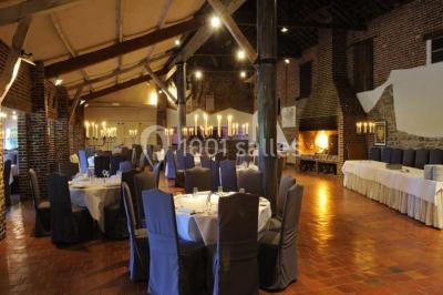 Salle de réception avec tables dressées, nappes beiges, chandeliers et poutres apparentes sous un toit vitré.