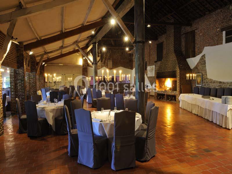 Salle de réception avec tables rondes dressées, chaises couvertes, cheminée allumée et éclairage chaleureux.