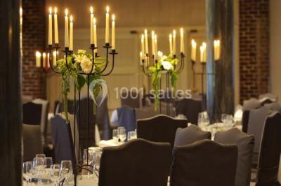 Salle de réception avec tables dressées, nappes beiges, chandeliers et poutres apparentes sous un toit vitré.