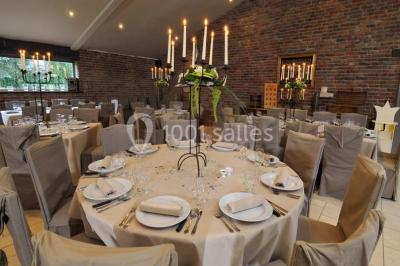 Salle de réception avec tables dressées, nappes beiges, chandeliers et poutres apparentes sous un toit vitré.