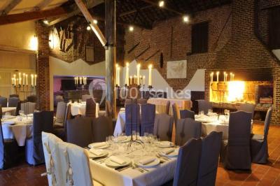 Salle de réception avec tables dressées, nappes beiges, chandeliers et poutres apparentes sous un toit vitré.