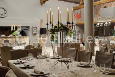 Salle de réception avec tables dressées, nappes beiges, chandeliers et poutres apparentes sous un toit vitré.