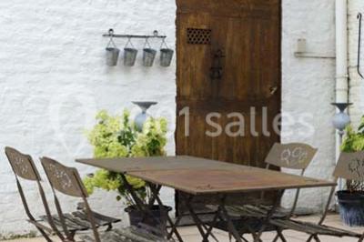 Table et chaises en métal sur une terrasse en dalles, devant un mur blanc avec une porte en bois et des plantes décoratives.