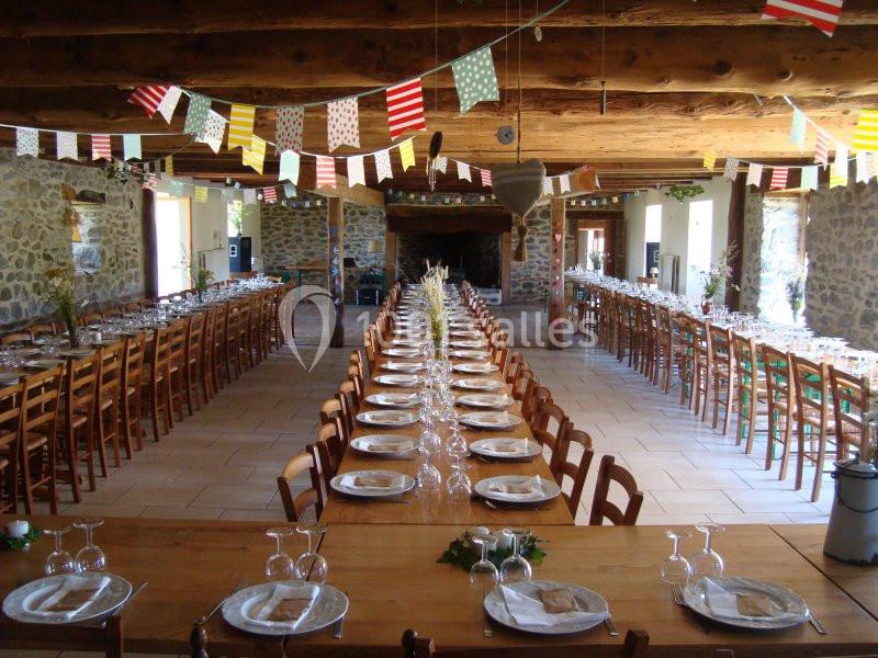 Grande salle de réception rustique avec longues tables dressées, décorée de guirlandes colorées sous un plafond en bois.