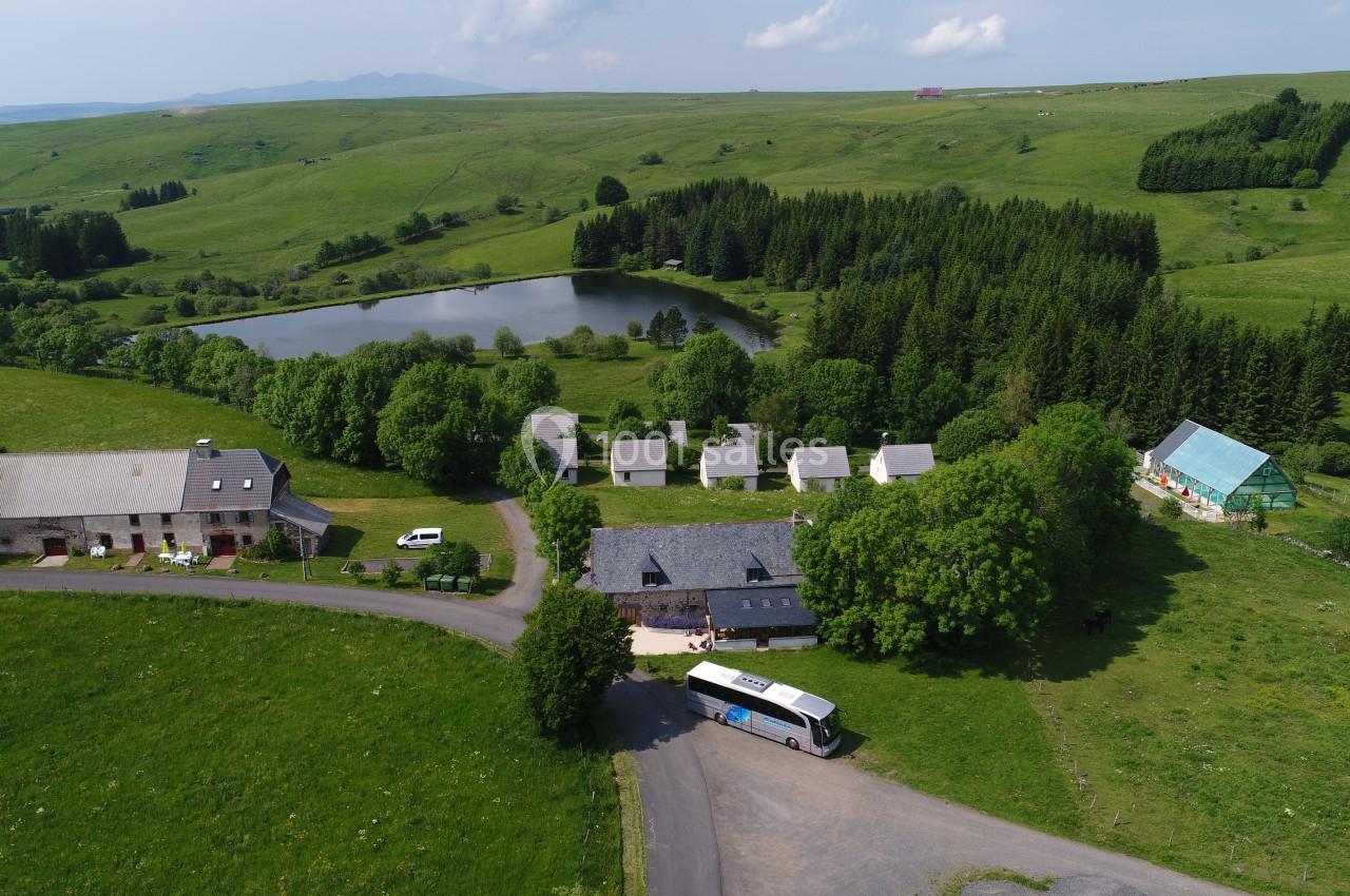 Vue aérienne d'un hameau entouré de prairies verdoyantes, d'un étang et de forêts, avec un bus stationné sur une route.